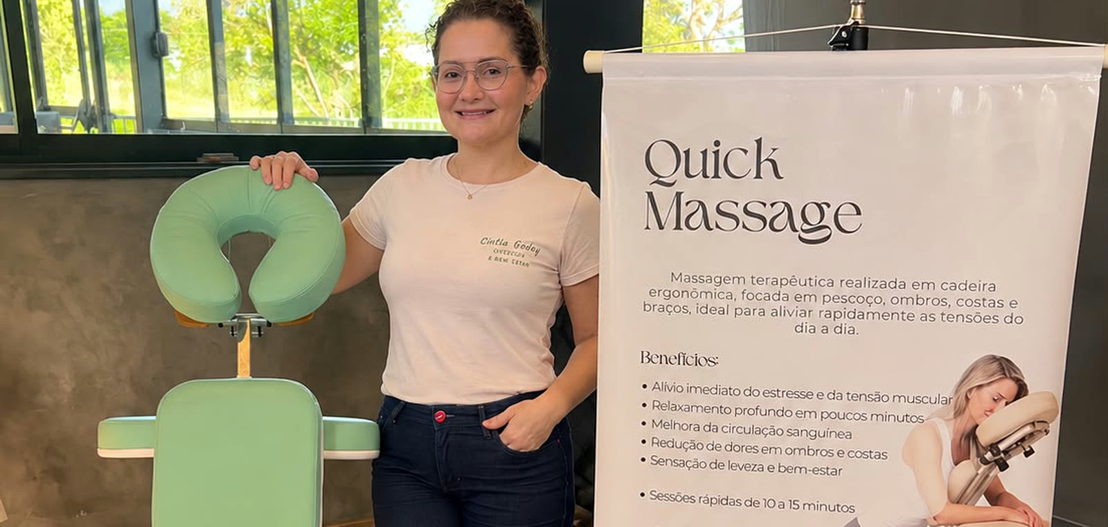 Cíntia Godoy ao lado da cadeira de quick massage e do banner do serviço